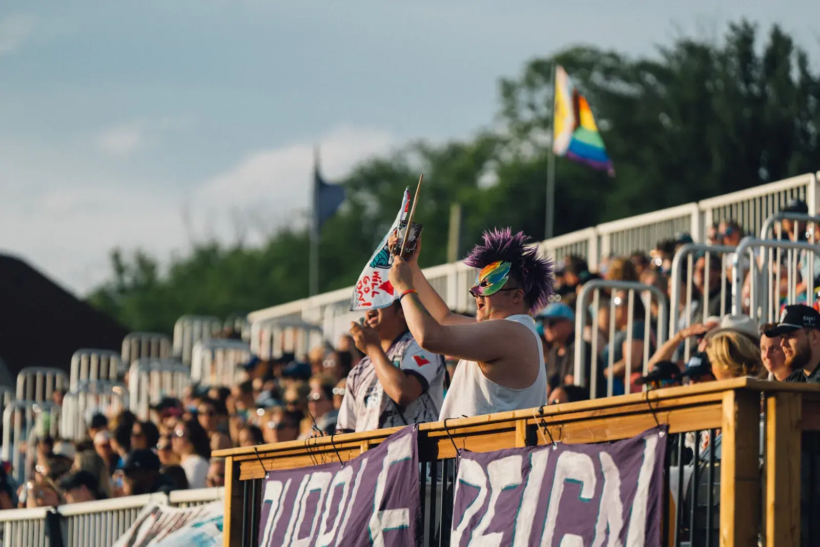Des partisans dans les gradins lors d’un événement estival, dont un portant un masque arc-en-ciel et agitant un drapeau